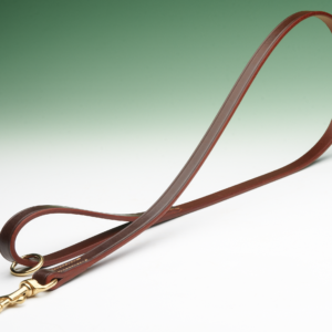 A brown leather dog lead lies curved on a white surface against a green backdrop. The leash has a brass snap hook and ring.