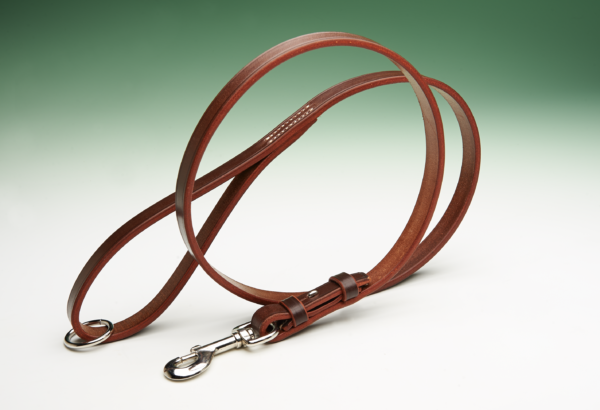 A brown leather dog lead, lying in a loop, rests on a white surface against a blurred green background. The leash has a silver hook at one end.