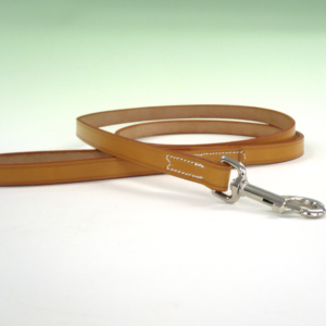 A light brown leather dog lead lies coiled on a white surface; a silver clasp is attached to one end. The leash shows subtle stitching details.